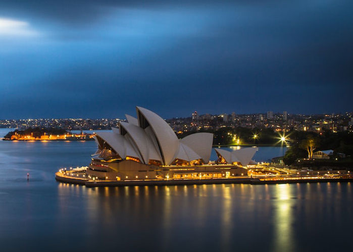 Evening view of the iconic Sydney Opera House in Australia, with its unique shell-like architecture illuminated against the dark, cloudy sky, surrounded by calm waters of Sydney Harbour and the cityscape in the background.