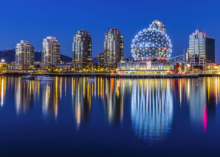 Nighttime cityscape of Vancouver, Canada, showcasing the illuminated Science World building with its iconic geodesic dome, reflecting beautifully on the calm waters of False Creek, surrounded by modern high-rise buildings.