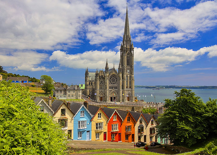 Colorful houses along a hillside with a view of a grand cathedral and waterfront in Cobh, Ireland, under a vibrant blue sky with scattered clouds