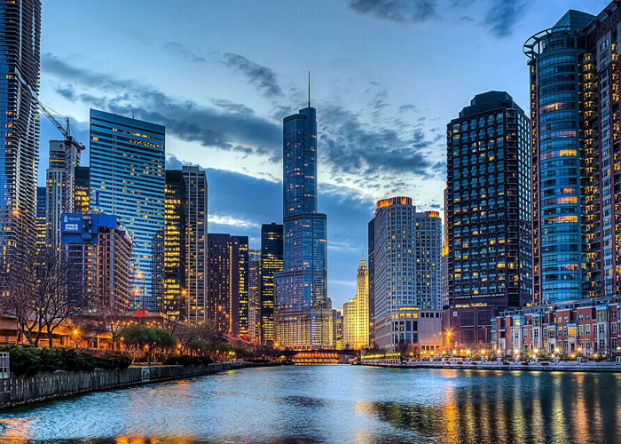 Evening cityscape of Chicago, USA, featuring the illuminated high-rise buildings along the Chicago River, with the iconic Trump Tower standing tall in the center, reflecting against the calm river water under a cloudy sky.